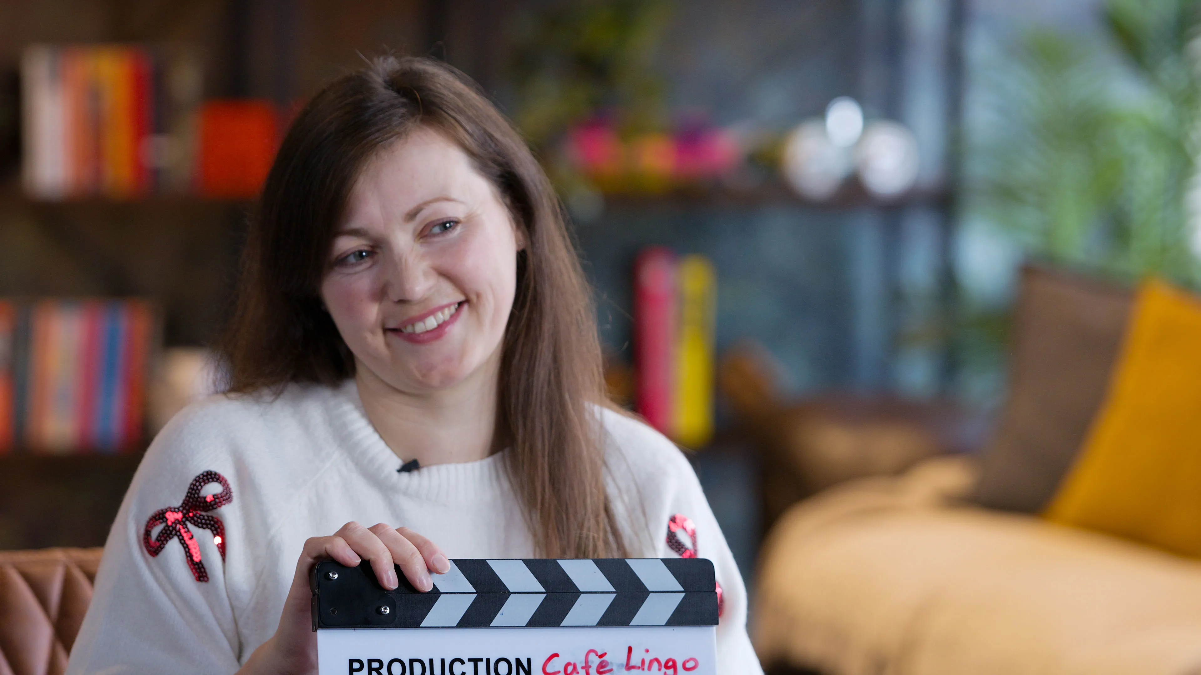 Smiling woman in a white sweater with bow designs holds a clapperboard labeled "Production: Cafe Lingo" in a cozy, colourful living room setting.